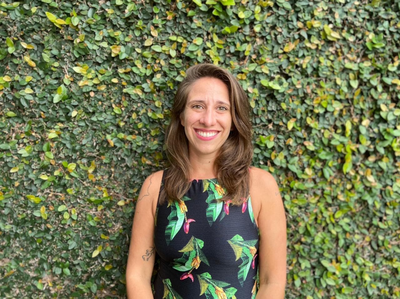 Carolina is a white women with long brown hair wearing a black top with leaves on it. She stands in front of a large green bush that fills the entire frame.